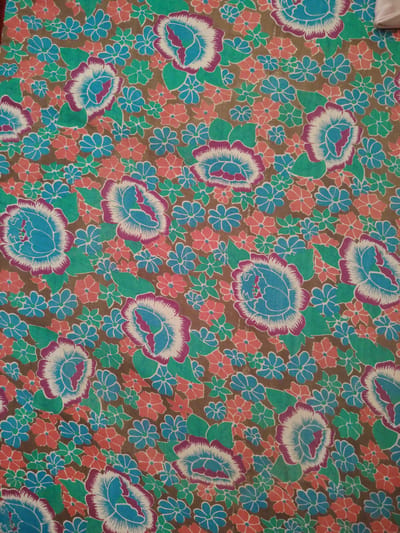 The image displays a close-up, flat lay view of a fabric with a floral pattern. The fabric has a brown background, densely covered with various flowers and leaves. The flowers are depicted in shades of blue, pink, and white, with some having purple outlines. The leaves are rendered in green. There are larger, more detailed flowers with blue centers, white petals edged in purple, and green leaves, as well as smaller, simpler flowers in blue, pink, and teal. The pattern is repeated across the entire visible surface. No people or specific environmental context of Pitnak, Uzbekistan are present in this image.
