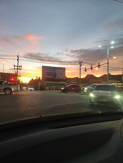 This media file captures a street scene in Khlong Thom, Thailand, during what appears to be sunset or dusk. The image is oriented vertically, with the sky occupying the left side and the street and roadside on the right.

The sky displays a gradient of colors, transitioning from a soft blue at the top to warm oranges and pinks near the horizon, indicating the sun is setting or has recently set. Wispy clouds are visible, catching the fading sunlight. Several streetlights are illuminated, casting a bright glow.

On the right side of the frame, a street is lined with various elements. Cars are present, suggesting ongoing traffic. A prominent billboard, with Thai text, is visible, advertising "ทูเคสปอร์ต" (Two Case Sport) and mentioning "กว่า 1,000 ทีมไว้ใจเรา" (Over 1,000 teams trust us). Other signs and shopfronts can be seen further down the street, including one with "Chang" branding and another reading "ร้านโกชาย" (Ko Chai Shop). Traffic lights are visible, with red lights illuminated, suggesting a stop. Power lines and utility poles are also a notable feature of the streetscape.

The overall impression is that of a bustling urban environment at the end of the day, with the natural beauty of the sunset contrasting with the man-made elements of the street. The lighting suggests it's twilight, with artificial lights beginning to dominate as natural light fades.