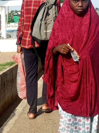 The image depicts two individuals on a sunlit, outdoor walkway. The person on the left wears a red and black plaid long-sleeved shirt, dark pants, and sandals, carrying a pink and white patterned bag. They have a grey backpack with the "Microsoft T" logo visible on it. The person on the right is wearing a striking red hijab with a floral pattern and a white and grey patterned skirt. They are holding out a few banknotes, suggesting a transaction or offering. The scene appears to be set in Garko, Nigeria, with a backdrop of greenery and a white structure. The lighting suggests it is daytime, likely morning or afternoon, with clear weather. The individuals' postures are natural, with the person on the right appearing to be engaged in an interaction while the person on the left walks alongside them.