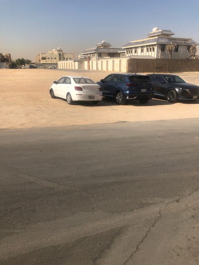 A row of parked cars, including a white sedan, a dark-blue SUV, and a black vehicle, lines a sandy patch beside a road. Buildings, some with distinct architectural features, are visible in the background against a bright sky. The road surface exhibits cracks and signs of wear.
