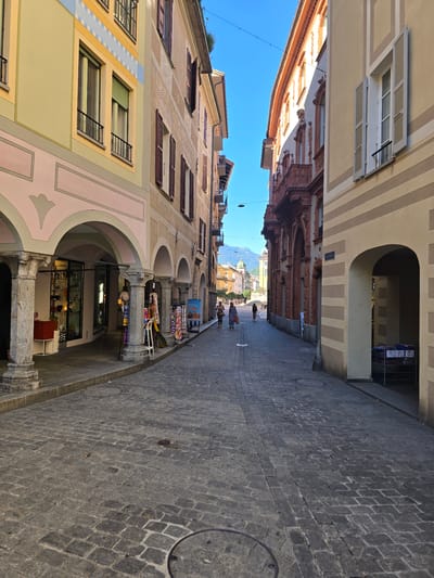 This image depicts a street scene in Bellinzona, Switzerland, under a clear blue sky, suggesting a sunny day. The perspective is from a low angle, looking up and along a narrow street flanked by historic buildings.
The buildings on either side of the street exhibit varied architectural styles and colors. The building on the left features pastel hues of yellow, pink, and beige, with multiple windows, some adorned with dark shutters. The building on the right displays a warm terracotta color with ornate architectural details and a series of arched walkways at ground level. The street itself is paved with dark cobblestones.
In the distance, mountains are visible, hinting at the mountainous landscape of Switzerland. Several people are scattered along the street, some walking and others appearing to be browsing or congregating. Objects visible include storefront displays with colorful textiles, outdoor furniture, and shopping carts. One building features a large arched entrance. The overall impression is of a vibrant, pedestrian-friendly urban environment, possibly a marketplace or shopping area.
No visible text is discernible in the image. The time of day appears to be daytime, given the bright sunlight and shadows cast. The scene evokes a sense of everyday life and commerce in a historic European town.