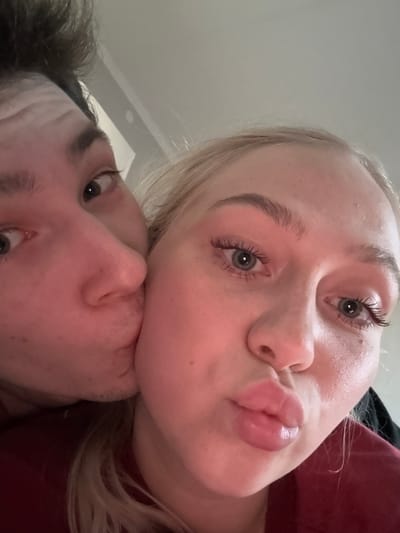 This image depicts two individuals in a close-up indoor setting within the city of Yanino-1, Russia. A male, visible from the left side of the frame, is shown with dark hair, dark eyes, and light stubble on his chin and jawline. He is actively kissing the left cheek of a female positioned predominantly on the right and center of the frame. The female has blonde hair, blue-grey eyes, and discernible eyelashes, possibly enhanced with mascara. Her facial expression is characterized by pursed lips, forming a pout, and she appears to be looking directly into the camera. Her skin is fair with minor freckles visible. Both individuals are wearing dark-colored upper garments; the female's garment appears to be a dark red or maroon. The background is an undifferentiated, light-colored surface, suggesting a simple wall or ceiling.
