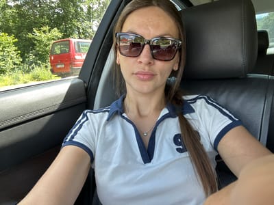 A woman with long brown hair, wearing a white polo shirt with blue trim and the number '9' on it, and sunglasses, sits in the driver's seat of a car. Her left arm is visible, resting on the car's interior. Outside the car window, a red van is parked on the side of a road, with lush green trees and foliage in the background. The setting appears to be a rural or suburban area, possibly near Dulovine, Montenegro, given the presence of green vegetation and a paved road. The lighting suggests it is daytime, with sunlight casting shadows.
