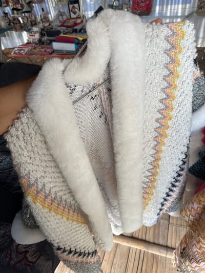 This is a close-up, eye-level shot of a person's arm and torso holding a fluffy white-trimmed sweater with a zig-zag pattern of pink, yellow, and gray. The sweater is hanging on a metal rack in what appears to be an outdoor market stall in Mai Chau, Vietnam. In the blurred background, other textiles, bags, and market goods are visible, suggesting a vibrant commercial setting. The wooden plank floor and the natural light indicate an authentic, possibly rural, market environment.