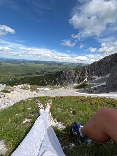 The image shows a person's legs and feet resting in the foreground, positioned on a grassy slope with scattered rocks and patches of snow. The person is wearing light gray sweatpants and leopard-print sneakers on their left foot, and a blue sneaker with white accents on their right foot. To the left of the foreground, a rocky slope descends into a wide valley. The valley features green fields, scattered buildings, a small lake, and rolling hills in the distance. A ski lift structure is visible in the mid-ground on the left side of the slope. The background is dominated by a vast blue sky with scattered white clouds. The rocky mountain to the right of the frame shows evidence of snow accumulation in crevices. The location is identified as Pašina voda, Montenegro. The overall scene depicts a scenic mountain landscape under a bright, cloudy sky.
