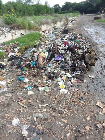 The image shows a large pile of garbage and debris in a ditch along a dirt road in Athi River, Kenya. The garbage is mostly composed of plastic bags, cardboard boxes, and other miscellaneous trash. There is also a small amount of water in the ditch. The area appears to be overgrown with vegetation. A small concrete wall and green vegetation can be seen in the background, as well as a dirt road. The image was taken in the morning and is likely a result of poor waste management in the area.