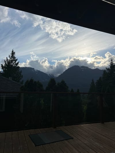 The photograph captures a scene in Squamish, Canada, showcasing a view of mountains framed by a deck or balcony setting. The sky is filled with white, streaky clouds against a blue backdrop, suggesting a partly cloudy day. A large mountain range dominates the background, partially obscured by the clouds. In the midground, trees add to the natural scenery. The foreground includes a wooden deck area, with a black mat or rug visible. To the left, a part of a building structure is present, possibly an overhang or roof. The image suggests a moment of observing the landscape from a residential or lodging setting in Squamish.