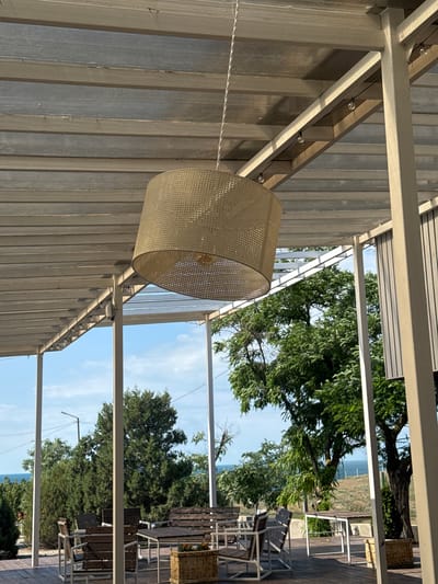 The image depicts an outdoor seating area with a canopy, likely a cafe or restaurant, in Sudak, Ukraine. The canopy is constructed from translucent corrugated panels supported by white metal beams and posts. A large, woven lampshade hangs from the center of the canopy. String lights are visible along the beams.
Below the canopy, there are several wooden tables and chairs with white metal frames, arranged on a wooden deck. In the background, lush green trees and vegetation are visible, with a glimpse of the sea and a blue sky. The overall scene suggests a pleasant outdoor dining environment.