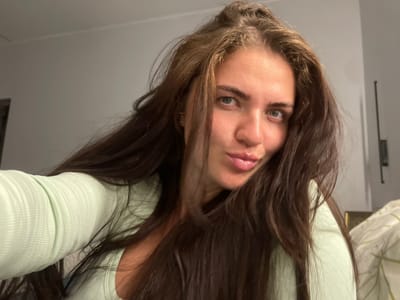 The image shows a close-up, selfie-style portrait of a young woman with long, dark brown hair, light blue eyes, and fair skin. She is wearing a light green, ribbed, long-sleeved top. Her lips are slightly parted in a pout, and her gaze is directed towards the camera. The background appears to be an indoor setting, with a plain gray wall and a glimpse of a patterned duvet or pillow on the right side of the frame. There are no visible elements that suggest a specific location within Мосметаллоконструкция Онт, Russia.
