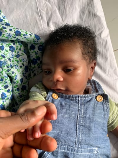 A baby lies on a floral-patterned sheet, wearing blue denim overalls and a striped shirt. An adult hand holds the baby's hand. The environment appears to be indoors, potentially a home. The location is Ibasa, Nigeria.