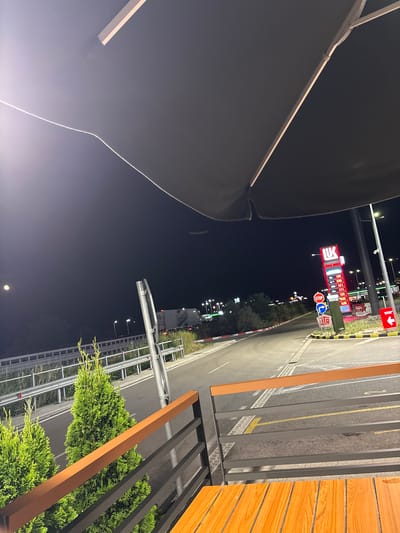 The image shows an outdoor seating area at night, likely at a gas station or roadside rest stop, with a dark canopy overhead. In the foreground, there is a wooden table and a railing. To the left, two small, well-maintained evergreen trees are visible. Beyond the railing, a paved road curves to the right, illuminated by streetlights. On the right side of the road, a LUKOIL gas station sign displays fuel prices, and various road signs, including a "STOP" sign, are present. A white barrier with red and white reflective strips runs alongside the road. In the background, there are more lights, suggesting a commercial area or highway. The sky is dark, and a faint light, possibly the moon, is visible in the upper left corner. The scene appears to be located in Chuchuligovo, Bulgaria. There are no people visible in the image.