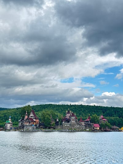 The image depicts a lakeside view in Satka, Russia, on a cloudy day, likely in the late afternoon. The scene features a row of rustic, fantasy-themed buildings nestled along the shoreline, backed by a dense, dark green forest. These structures have distinctive red-tiled roofs and appear to be made of stone and wood, resembling a medieval or fairytale village. Several buildings have turret-like elements, adding to the whimsical aesthetic.

The water in the foreground is rippling, suggesting a gentle breeze. The sky is a mix of dark, heavy clouds and patches of bright blue sky, indicating dynamic weather. One of the buildings has a bright yellow and blue object on its roof, possibly an umbrella or decoration. A small, dark bird is visible soaring in the distance against the cloudy sky.

The overall atmosphere is one of natural beauty blended with a touch of fantasy, creating a unique and intriguing landscape. No text is visible in the image.