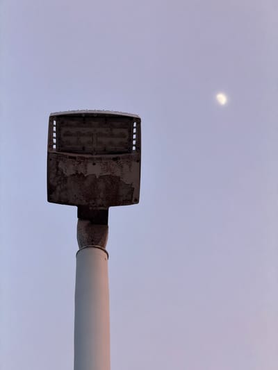 An upward shot of a street lamp post against a pale sky. The street lamp's housing is partially open, revealing what appear to be LED components. The housing is weathered and stained with rust. The pole of the street lamp is smooth and white, with a metal connector to the lamp housing that also appears weathered. A faint crescent moon is visible in the upper right quadrant of the sky. The scene is likely located in Miami Beach, United States of America, as indicated by the presence of a street lamp in a coastal urban environment.