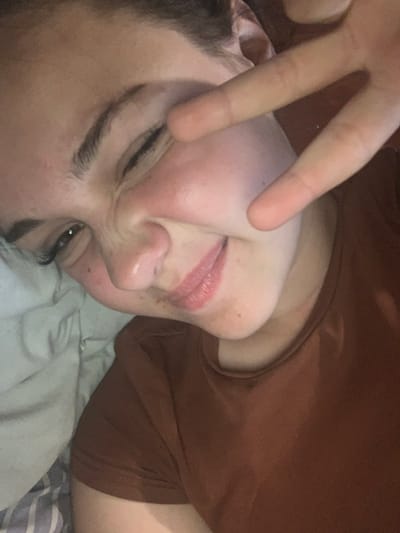 A close-up, slightly blurry selfie shows a person's face with a peace sign gesture over their eye. The person is winking and making a playful expression, with their mouth slightly open and lips colored. The skin appears fair with some blush on the cheeks and a few freckles. The background shows a white pillow and a brown shirt. There is no indication of the location being Hoholeve, Ukraine.