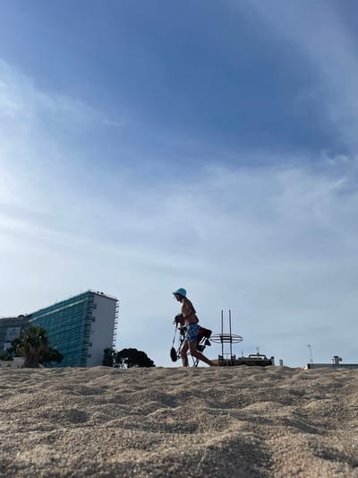 A person walks across a sandy beach in Canet de Mar, Spain. The person is wearing a blue hat and blue patterned shorts. They are carrying a pole with a dark object attached to it. In the background, a large building is under construction, covered in scaffolding and blue netting. Several trees are visible between the beach and the building. The sky is blue with some wispy clouds. The foreground is dominated by a close-up view of the sand.
