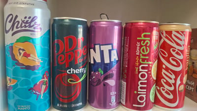 The image shows five beverage cans lined up on a shelf. From left to right, the cans are:
1. A white and blue can with "Chils SODA" written on it, featuring an illustration of a person lounging on a peach. The text "ВОТ ЭТО ОТКРЫТИЕ" and "СО ВКУСОМ МАНГО" is also visible.
2. A dark green can with "Dr Pepper cherry" written in red and white lettering.
3. A purple can with "FANTA" written in large white letters and an illustration of grapes. Text at the bottom indicates "0,33 л".
4. A pink can with "Laimon Fresh" written in green lettering, featuring illustrations of lime and lemon slices. Text indicates "LIME LEMON BERRIES" and "100% NATURAL INGREDIENTS".
5. A red can with the "Coca-Cola" logo in white lettering and "vanilla" written vertically.
The overall scene is clean and well-lit, suggesting a retail or home display environment. The cans are all upright and appear to be full. There are no people visible in the image. The background is a neutral gray, and the cans are positioned against what appears to be a light-colored surface. The image is focused on the beverage cans, with no extraneous details that would suggest a specific location beyond a place where such products are stored or displayed. No information is present in the image to indicate a location in the city of Stowbtsy, Belarus.