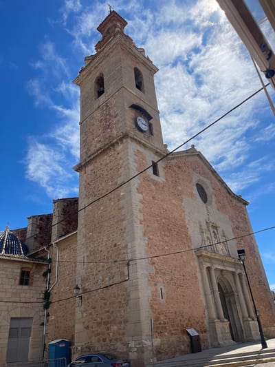 The image shows a low-angle view of the Església de la Mare de Déu del Roser (Church of the Virgin of the Rosary) in Puçol, Spain. The church is constructed of stone and features a clock tower. The sky is blue with scattered clouds. A partial view of a car and some street elements are visible to the right of the church.