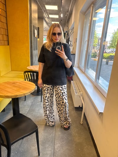 The image shows a vertical, full-body mirror selfie of a woman in what appears to be a cafe or restaurant. The woman has blonde hair, is wearing sunglasses, a black t-shirt, and leopard-print pants. She is holding a smartphone and taking the picture. She is standing next to a yellow booth and two round wooden tables. In the background, there is a large window with a view of trees and buildings, suggesting an urban setting. There is also a white radiator visible along the window sill, and what looks like a black chair in the foreground. The lighting suggests it is daytime, possibly late morning or afternoon. The overall mood is casual and self-aware. No visible text is clearly legible, except for a small logo on one of the wooden tables.