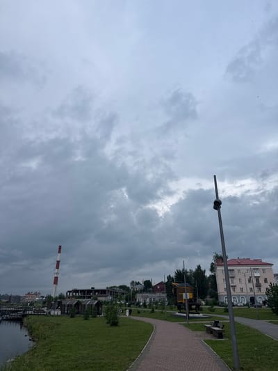 The image depicts an overcast day in the city of Lysva, Russia, likely near the waterfront. In the foreground, a brick pathway curves through a grassy area with several wooden benches. A tall, dark lamppost stands to the right of the path. Further back, a yellow and brown historical railroad car monument is visible. Beyond that, several small wooden structures are arranged on the grass. To the left, a body of water with wooden piers borders the grassy area. In the middle ground, a tall, red and white striped industrial chimney dominates the skyline, along with the skeletal remains of a large, unfinished building. Further in the distance, a multi-story apartment building with a red roof can be seen, along with other urban structures and trees. Two individuals are walking on the pathway in the middle ground. The sky is filled with heavy, grey clouds.