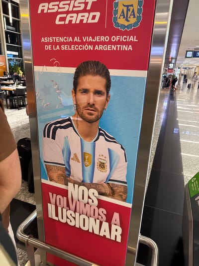 This media file captures a vertical promotional banner at Aeropuerto Internacional Ezeiza, Argentina. The banner features a close-up of a man, likely a soccer player, wearing an Argentina national team jersey. His gaze is directed forward, with a neutral expression. He has tattoos visible on his arms and a small earring. Text on the banner reads "ASSIST CARD" and "ASISTENCIA AL VIAJERO OFICIAL DE LA SELECCIÓN ARGENTINA" which translates to "Official Traveler Assistance of the Argentine National Team." Another prominent text in large, bold font says "NOS VOLVIMOS A ILUSIONAR," meaning "We got excited again." The background of the banner shows a light blue pattern and some celebratory streamers.

The setting appears to be an indoor public space, possibly an airport terminal, with glimpses of other people and luggage in the blurred background, suggesting travel activity. The lighting indicates daytime. A recycling bin with labels in Spanish and English ("Vidrio Glass-Vidro", "Plástico-Plastic", "Papel Paper") is visible in the upper right corner, further reinforcing the public facility context. The overall impression is one of promotion and support for the Argentine national team, likely tied to a travel assistance service.