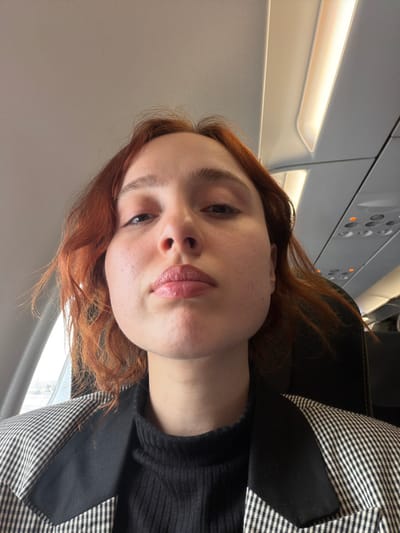 A young female with reddish-brown, shoulder-length hair is depicted in a close-up, upward-angled selfie taken inside a commercial airplane cabin. She has fair skin with visible pores, light brown eyebrows, and dark irises. Her lips are full and coated with a light pink or nude gloss, featuring a minor indentation on the upper lip. She wears a black ribbed turtleneck top underneath a black and white houndstooth or plaid blazer with solid black lapels.

Behind her, the dark headrest of a passenger seat is visible. To her left, a partial view through an aircraft window shows an overcast or hazy sky. Above her, the light grey airplane ceiling features a warm yellow-illuminated overhead lighting strip and a panel with multiple small, circular indicator lights, some of which are illuminated orange or red. The environment indicates the subject is aboard a commercial aircraft, in transit. Regarding the specified location of "Tuzla, Turkey," the aircraft is either at or in the vicinity of Sabiha Gökçen Airport (SAW), which serves the Istanbul area and is geographically close to Istanbul's Tuzla district, or within Turkish airspace.