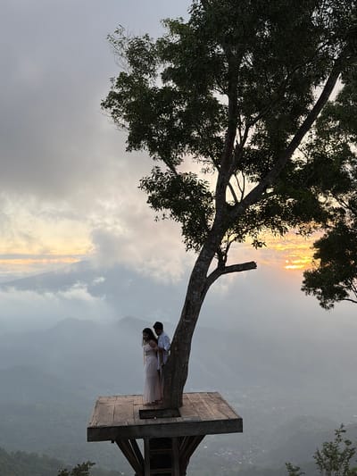 A couple is embracing on a wooden platform extending from a large tree. The man, wearing a white shirt, has his arms around the woman, who is in a long, light-colored dress. They are positioned on the platform, which is elevated high above a misty landscape of rolling hills and a distant village. The scene is framed by the branches of the tree, with the sun setting or rising behind a cloudy sky in the background, casting a warm glow. The location is identified as Abang, Indonesia.
