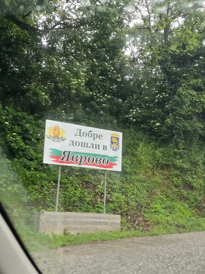 A street sign in Yavrovo, Bulgaria, reads "Добре дошли в Яврово" (Welcome to Yavrovo). The sign features the Bulgarian coat of arms and the national flag colors of white, green, and red. It is mounted on two metal posts. The sign is positioned on the right side of a paved road, with a concrete barrier to its left. The surrounding environment is lush and green, with dense trees and vegetation. The road surface appears wet, possibly from rain. The image is taken from a vehicle, as indicated by the blurry foreground and reflections on what appears to be a car windshield.