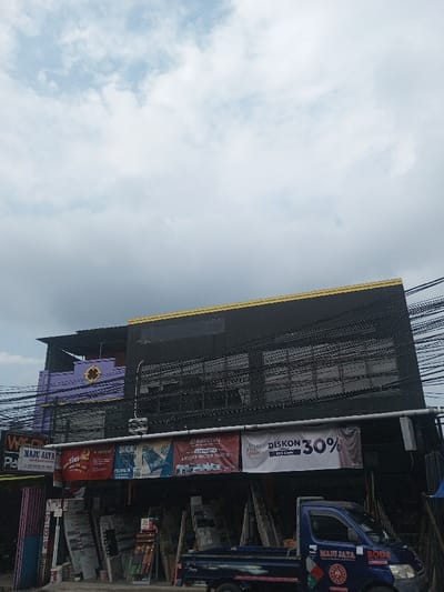 A two-story building with a black facade and yellow trim is seen under a cloudy sky. The second story has windows and power lines crisscrossing in front. Beneath the building are various businesses with signs for "VICON," "MAJU JAYA," "Tiles-lic," "BANDAL," and "ARISTON," advertising tiles, water heaters, and a 30% discount. A blue truck with "MAJU JAYA" branding is parked in front, suggesting a construction or home improvement supply store. A purple building with a sunflower emblem peeks out to the left.