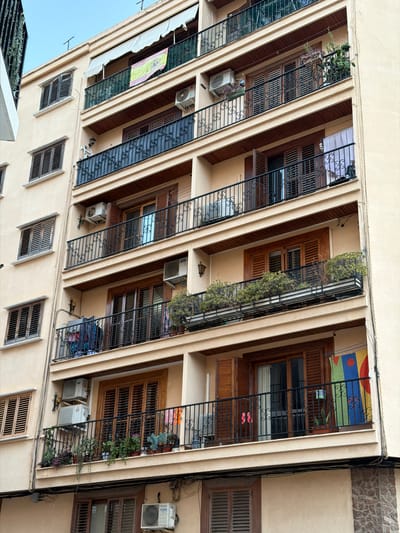 The image shows a facade of a multi-story apartment building. The building has a light beige exterior and features multiple balconies with black wrought-iron railings. Each balcony has wooden shutters on the windows. There are several air conditioning units mounted on the exterior walls. Various items are visible on the balconies, including plants in pots and hanging baskets, laundry hanging on a line, and some folded items. A Spanish flag is prominently displayed on an awning above one of the balconies. The building is situated in an urban environment, as suggested by the view of the sky and the architecture. The specific location is Benimàmet-beniferri, Spain.