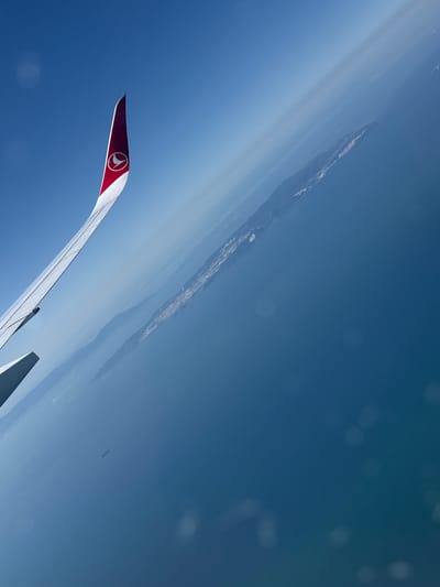 An aerial view from an airplane window shows a wingtip with a Turkish Airlines logo against a clear blue sky. Below the wing, the sea stretches out to a distant coastline, possibly a city, with visible lights and structures. A small ship is visible in the water. The image is tilted, suggesting the aircraft is banking.