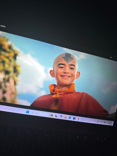 The image displays a laptop screen, rotated clockwise, showcasing a scene from a live-action adaptation of Avatar: The Last Airbender. The central subject is a young boy portraying Aang, characterized by his bald head and the distinctive blue arrow tattoo on his forehead. He wears a traditional orange and red outfit, consistent with Air Nomad attire, and is captured in a cheerful pose, smiling broadly with a happy expression while looking slightly upwards and off-screen.
The background visible on the screen is an outdoor setting under a clear, bright blue sky with soft white clouds. Blurred green foliage in the lower portion suggests a natural environment, possibly a hill or forest. The laptop's Windows taskbar is prominent along the right edge of the screen, displaying the time "8:26 PM" and the date "4/21/2025". Several application icons are visible, including those for Microsoft Edge, File Explorer, Mail, Photos, and Netflix. A search bar with the text "Search" is also present. The word "Netflix" is also visible, rotated, in the bottom left corner of the screen. A section of the laptop's black keyboard is partially visible to the right of the screen. The screen itself shows signs of use, with visible dust, smudges, and fine scratches. This image was captured in Kirana, Nigeria.