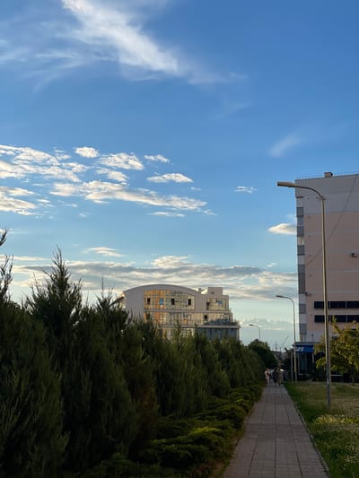 In Rozsoshentsi, Ukraine, an outdoor scene unfolds under a vast blue sky dotted with scattered white clouds. A paved walkway, constructed from rectangular stones, guides the viewer's gaze into the distance. To the left of the walkway, a dense row of dark green evergreen bushes forms a lush barrier. On the right side of the walkway, a patch of green grass is interspersed with numerous small white wildflowers.
Further down the walkway, a group of three individuals is walking away from the camera. They are positioned in the middle ground of the image.
In the background, two modern buildings stand out. The building on the left features a curved facade with numerous windows reflecting the sky and surrounding environment. The building on the right is taller and displays a lighter colored exterior with visible windows and what appear to be solar panels on its lower section. Two streetlights are visible along the walkway, one closer to the viewer and another further down. In the far distance, between the buildings, industrial cranes are faintly visible against the horizon. The overall lighting suggests it is either late afternoon or early evening.