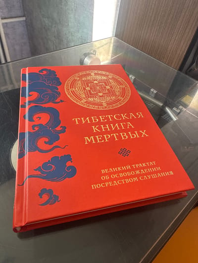A red book with gold lettering and blue cloud designs is placed on a glass surface. The book is open to a page with a circular, mandala-like design in gold. The title of the book appears to be "Тибетская Книга Мертвых" (Tibetan Book of the Dead). The environment appears to be indoors, possibly a home, with a record player visible in the background. There are no people or animals present in the image.