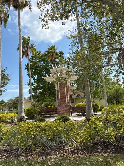 The image captures a serene outdoor scene in Parrish, United States, featuring a prominent brick and white pergola structure with benches situated beneath it. The pergola is surrounded by lush greenery, including tall palm trees and dense bushes with yellow and green foliage. A building with a light yellow facade and a brown roof is visible in the background, partially obscured by trees. The sky above is a vibrant blue with scattered white clouds, suggesting a bright, sunny day. The ground is covered with grass and fallen leaves, adding to the natural ambiance. There are no people or animals present in the image. The overall impression is one of a well-maintained park or community gathering space, offering a tranquil spot for relaxation.
