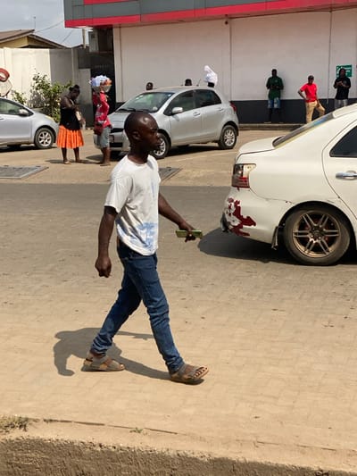 The image depicts an outdoor commercial or street environment in Ablekuma, Ghana, under direct sunlight. A dark-skinned male, with a shaven head, walks from left to right across the mid-ground. He wears a white t-shirt with faint blue graphics, blue jeans, and brown open-toed sandals, holding a green-cased smartphone in his right hand. His shadow is cast to his left, indicating direct overhead sun.

In the immediate background, a white sedan is parked, showing significant damage to its rear driver's side, including missing sections of the bumper, exposed red undercoat or paint transfer, and a broken or absent taillight. It is equipped with multi-spoke bronze wheels. To the left, a silver compact hatchback is parked. Adjacent to it, a dark-skinned female, dressed in a black top and an orange pleated skirt, stands looking downwards. Behind her, another dark-skinned individual, wearing a plaid shirt and light shorts, balances a large, round, laden basket on their head. A white cloth object is draped over the roof of the silver hatchback.

The far background features a cream-colored commercial building with a prominent red fascia. Along its wall, three dark-skinned males are standing: one in a dark green t-shirt and dark shorts, one in a red t-shirt and tan pants leaning against the wall, and another in a dark t-shirt and dark shorts observing a phone. The ground is paved with light-colored material, and a concrete curb defines the immediate foreground.