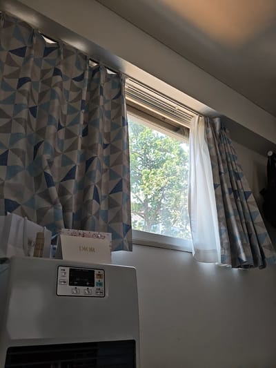 The image depicts an indoor scene, likely a room, in Sapporo, Japan, as indicated by the context of the request. The primary focus is on a window with curtains. On the left, a patterned curtain with geometric shapes in blue and gray is partially drawn. To the right of it, a sheer white curtain is visible, and further to the right, another patterned curtain, identical to the one on the left, is also drawn. Through the window, the lush green foliage of a tree is visible, suggesting an outdoor environment with abundant plant life.
In the foreground, on top of a white appliance that appears to be a heater, several items are placed. These include two white shopping bags, one with "DIO" visible on it, a small bottle with a gold cap, and a white box with the "DIOR" logo printed in gold. The appliance itself has a control panel with various buttons and a digital display, indicating it is an electronic device. The wall behind the objects and the window is painted a light, neutral color. The lighting suggests it is daytime, with sunlight filtering through the window. There are no people visible in the image, and no actions or interactions are depicted. The scene is static and appears to be a still life arrangement in a domestic or commercial setting.