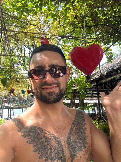 A man with a beard and sunglasses is posing for a selfie outdoors in Ninh Ích, Vietnam. He is shirtless and has a large tattoo of wings on his chest. He is standing under a tree with yellow flowers and decorative heart-shaped ornaments hanging from the branches. In the background, there is a thatched-roof building and the ocean. The man is smiling and has his left hand raised.