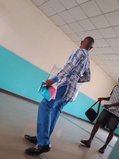 The image shows two men in an indoor location in Umueshi, Nigeria. The man in the foreground is wearing a patterned long-sleeved shirt, blue pants, and black shoes. He is holding papers. The man in the background is wearing a patterned t-shirt, dark shorts, and sandals. He is carrying a black bag. The room has blue and white walls and a tiled ceiling.