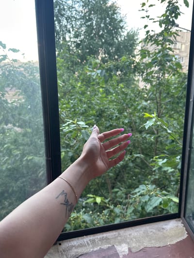 A person's arm and hand extend from the left side of the frame, reaching towards a window. The hand, with bright pink fingernails, is open and facing the viewer. A thin gold bracelet is visible on the wrist, and a black tattoo is present on the forearm. The scene appears to be an interior space looking out a window at a dense canopy of green trees and foliage. The window frame is dark and appears to be made of metal. Peeling paint or wallpaper is visible on the windowsill in the foreground. The image is captured from a medium-close shot, focusing on the arm and hand against the natural backdrop. The lighting suggests it is daytime. The location is specified as Magnitogorsk, Russia.