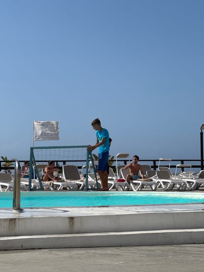 The image depicts a sunny outdoor scene at what appears to be a resort or hotel, likely the Saracen Hotel & Congress Centre in Capaci, Italy, given the flag. In the foreground, a young man in a blue t-shirt and blue shorts is standing next to a makeshift goal net with a blue frame, positioned at the edge of a turquoise swimming pool. Behind him, several white lounge chairs are occupied by people. To the left of the pool, a woman in a black bikini is seated on a lounge chair, holding a white object, possibly a drink or food. Further back, another person is visible sitting on a lounge chair. To the right, a man with a bare chest and blue shorts is seated on a lounge chair, holding a drink. A row of empty white lounge chairs stretches across the background, leading to a wooden fence and more distant palm trees under a clear blue sky. The area around the pool appears to be paved with concrete.