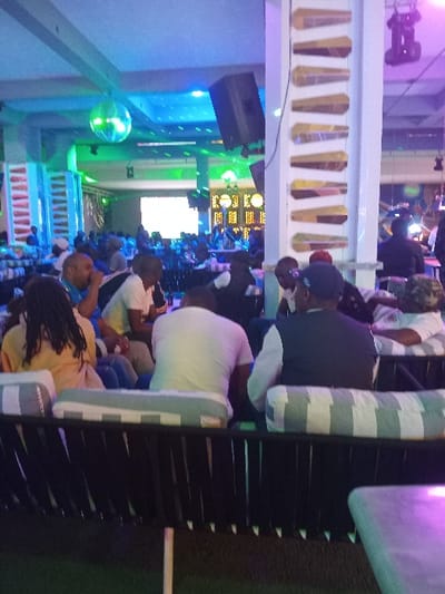 The image depicts the interior of a social establishment, likely a lounge or nightclub, located in Syokimau, Kenya. The scene is characterized by dynamic artificial lighting, predominantly in shades of blue and green, suggesting an evening or nighttime setting.

Multiple individuals, primarily adults, are seated in various arrangements across the venue. In the immediate foreground, the back of a dark-colored sofa with grey and white striped cushions is visible, occupied by at least three patrons: a person with dreadlocks wearing a light-colored garment, a male in a white t-shirt, and a male wearing a dark vest over a white long-sleeved shirt. A light-colored table is partially visible on the far right.

Midground observations reveal numerous other patrons seated at tables or on lounge-style seating, engaged in social interaction. A prominent white vertical pillar on the right side of the frame is decorated with a stacked arrangement of ten distinct, elongated, wooden or gourd-like objects, possibly stylized musical instruments or decorative elements.

In the background, the expansive interior features a disco ball suspended from the ceiling, reflecting ambient light. Large, dark-colored speakers are mounted from the ceiling structure. A large, bright rectangular screen or display is visible in the distance, emitting a white light. Additional decorative vertical elements are present on other pillars or walls. The flooring in the foreground appears dark, possibly artificial turf. The overall environment suggests a vibrant and active social venue.