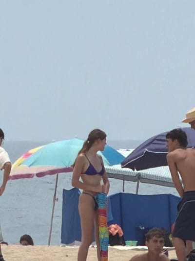 The image depicts a sunny day at the beach in Espinho, Portugal. In the foreground, a young woman in a blue bikini stands holding a colorful striped towel. To her right, a young man in dark swim trunks is partially visible from the side, his back to the camera. In the background, several beach umbrellas are set up, with one large umbrella displaying a mix of blue, yellow, and pink hues. Another dark blue umbrella is prominent behind the young man. The sandy beach stretches towards the ocean, where the water and horizon are visible under a hazy, light blue sky. Other beachgoers and beach structures are blurred in the distance, suggesting a busy atmosphere.