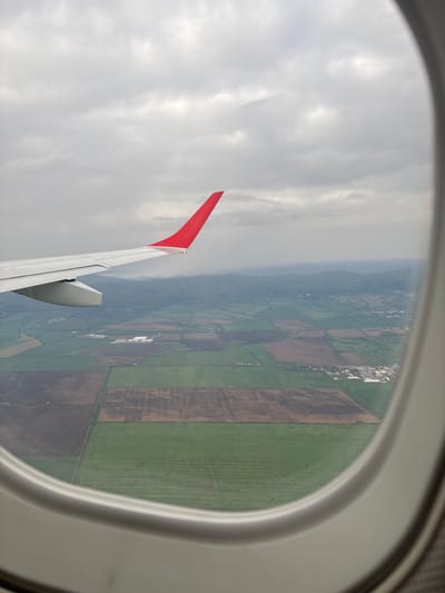 A view from an airplane window shows the wing of the plane with a red tip, outside is a landscape with fields of brown and green and a settlement with white buildings. The sky is overcast. This is likely taken near Schwechat, Austria, based on the landscape characteristics.