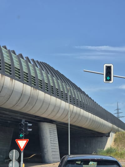 A wide-angle, eye-level outdoor shot captures a view of a modern overpass structure in Sanem, Luxembourg. The overpass is a curved, grey concrete barrier with an arched top section covered in slanted glass panels and dark grey louvers, suggesting a noise barrier. The sky above is a clear, bright blue with a few wispy white clouds.
In the foreground and mid-ground, traffic lights and signage are visible. A traffic light suspended from a pole displays a green arrow pointing left. Another traffic light, closer to the ground and mounted on a pole, also shows a green left arrow. Below this, a red and white triangular yield sign is prominently displayed. Further down the pole, two circular traffic signs with white backgrounds and grey markings are visible. The underside of the overpass is concrete, showing support pillars and a dark, cavernous space beneath.
To the right of the overpass, a tall, lattice-style electricity pylon stands against the blue sky. The ground near the overpass is covered with dry, yellowish-green grass and some greenery. The roof of a dark-colored car is visible at the very bottom of the frame, suggesting the photo was taken from inside a vehicle. The overall impression is one of urban infrastructure in a bright, clear, daytime environment.
