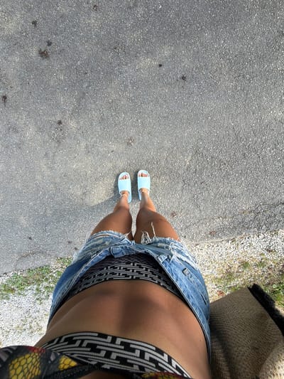 A person stands on a paved surface, looking down. They are wearing light blue slide sandals, a denim skirt with frayed edges, and patterned underwear. A portion of a straw bag is visible on the right side. The ground is asphalt with small stones and some dark spots. The scene appears to be outdoors, possibly in a park or along a street, in Chavei, Bulgaria.