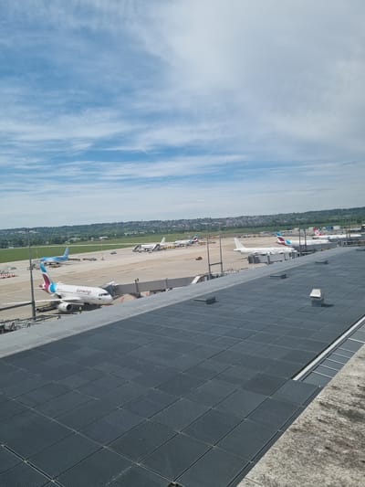 The image shows an airport scene in Echterdingen, Germany. Several airplanes are visible on the tarmac, including a Eurowings aircraft. The sky is partly cloudy. A building with a dark grey, patterned exterior wall is in the foreground.