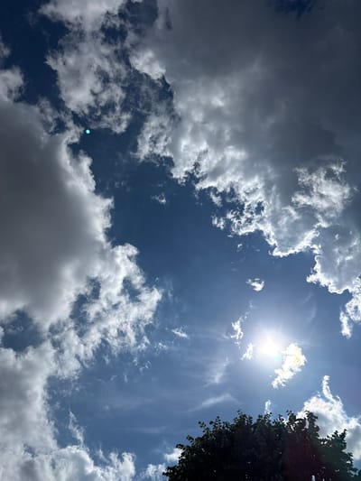This is a low-angle, full shot of a cloudy sky in Slavyani, Bulgaria. The sun is shining brightly through the clouds, creating a lens flare and a bright halo effect. The clouds are a mix of dark and light gray, with some white, fluffy clouds scattered throughout. In the bottom right corner of the image, the dark silhouette of a tree is visible, with its leaves obscuring a portion of the sky. The sky is a deep blue, indicating it is daytime. There are no people or animals visible in the image. The overall impression is of a dynamic and somewhat dramatic sky.