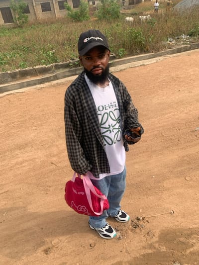 A male individual is observed standing on an unpaved, reddish-brown dirt road in Eika-Adagu New Layout, Nigeria. He presents with characteristics of dwarfism. The individual wears a black "Champion" baseball cap, a white t-shirt featuring a green "LOEWE" logo, an open dark-grey and white checkered long-sleeve shirt, light-blue jeans, and black, white, and light-blue sneakers. In his left hand, he holds a black smartphone. In his right hand, he carries a bright pink "Hello Kitty" themed bag with pink straps. His gaze is directed towards the camera, maintaining a neutral expression. To the immediate left, a low concrete curb borders an elevated area covered with dry, tall grass and weeds. In the background, several unfinished or dilapidated concrete block structures with dark openings are visible amidst the dry vegetation. Further in the distance, two indistinct figures, one human and one animal (possibly a goat or sheep), are discernible against the landscape. The scene is illuminated by natural daylight.