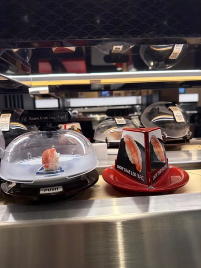 The image depicts a close-up, eye-level view of a conveyor belt sushi display at a restaurant, likely in Beaverton, Oregon, United States of America. Two sushi plates are prominently featured. The plate on the left is covered with a clear dome and contains a single piece of sushi, appearing to be crab or shrimp. A black label above the dome reads "(148) Snow Crab Leg (1-pc)". The plate on the right is red and is topped with a triangular display featuring images of snow crab legs and the text "SNOW CRAB LEG (1PC)" on each visible face. The background shows blurred shelves with more covered sushi plates, suggesting a busy sushi bar environment. The lighting is artificial, illuminating the sushi and the metallic surfaces of the display.