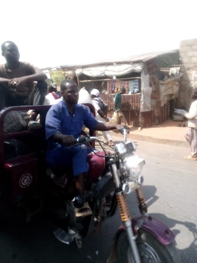 The image depicts a street scene in Tudun Murtala, Nigeria, under bright daylight conditions. In the foreground, a dark-skinned male operator, wearing a blue short-sleeved uniform, drives a dark red three-wheeled motorcycle. His hands are on the handlebars. Seated in the dark red passenger compartment directly behind the operator is a second dark-skinned male, wearing a brown t-shirt and dark trousers, looking towards the right of the frame. He has a ring on his right hand. The vehicle's front fender is purple, and its front suspension features exposed yellow/orange springs. A white circular emblem is visible on the side of the passenger compartment.
The background reveals a bustling urban environment. To the right, a dirt roadside borders a paved area where the vehicle is positioned. A series of makeshift commercial structures or stalls constructed from corrugated metal sheets and tarpaulins line the background. One structure features a green tarpaulin roof and a white fabric awning, displaying various goods. A yellow banner with partially visible text, "POS AND D..." is also visible. Several indistinct individuals are present in the background, including one person in a green shirt near the stalls and another in light-colored attire further to the right. The sky overhead appears bright and hazy.